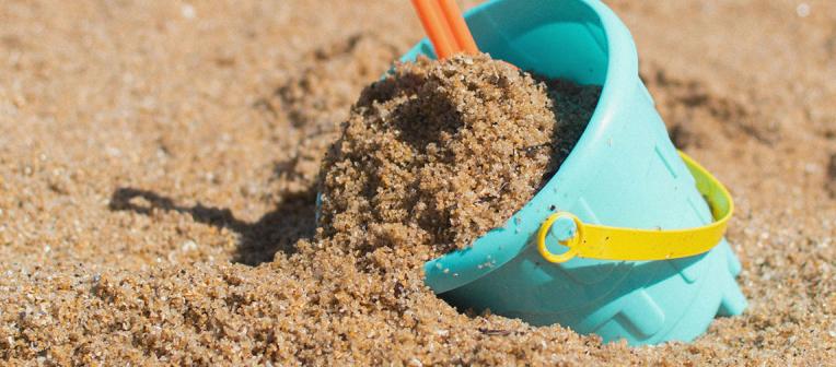 Closeup of a blue bucket with yellow handle full of sand being collected to build a sandcastle. Photo by Heather McKean on Unsplash